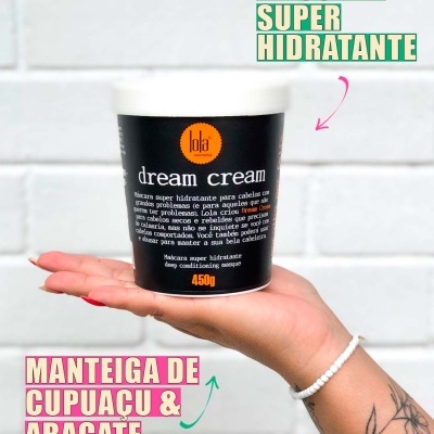 Frasco preto de máscara capilar hidratante dream cream da Lola segurado na mão com fundo branco tijolado