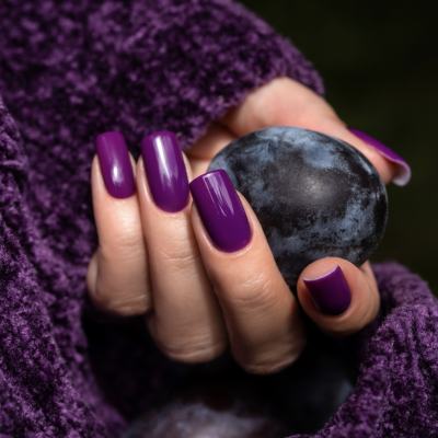 Mão feminina com unhas roxas brilhantes segurando ameixa com tecido roxo ao fundo.