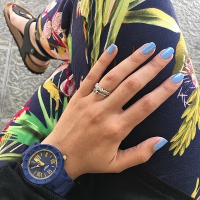 Relógio azul com mostrador redondo, anel prateado com pedras, calças florais coloridas e unhas azuis