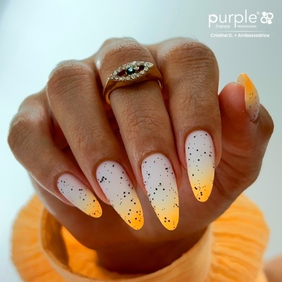 Mão com unhas decoradas em degradê branco para amarelo com manchas pretas e anel dourado com olho de pedrinhas