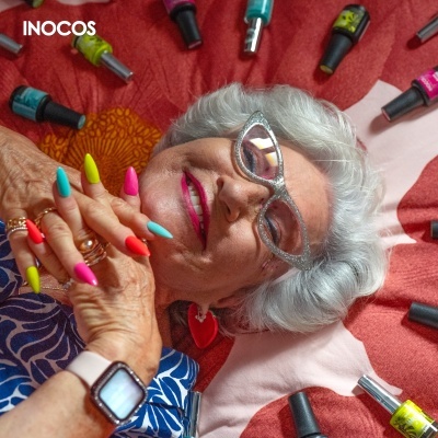 Mulher idosa com unhas coloridas neon, óculos brilhantes, rodeada por frascos de verniz INOCOS