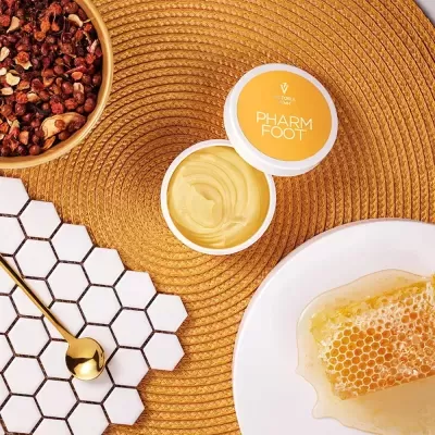 Frasco de creme para pés aberto ao lado de placa de mel, frutas secas e colher dourada sobre tapete mostarda.