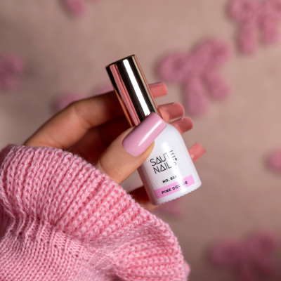 Frasco de verniz de unhas rosa claro SAUTE NAILS, sendo segurado por mão com unhas pintadas da mesma cor e suéter rosa.