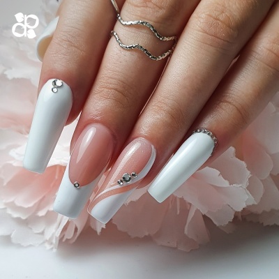 Unhas postiças decoradas com esmalte branco e rosa claro com brilhantes, anéis prateados no dedo e fundo de flores cor-de-rosa.
