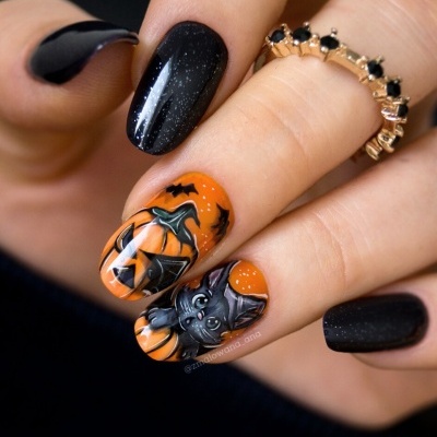 Unhas decoradas com tema de Halloween em preto e laranja com morcego e abóboras, mão com anel dourado com pedras pretas