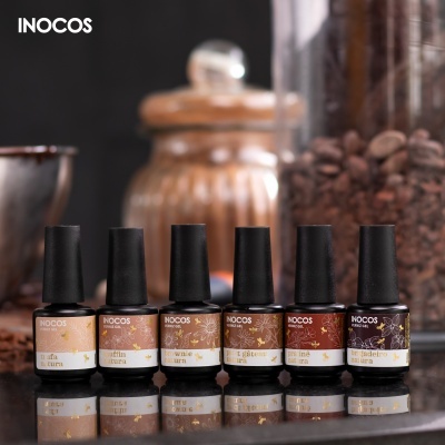 Seis frascos de verniz de unhas INOCOS alinhados em superfície reflexiva com fundo de grãos de café e frasco de vidro.