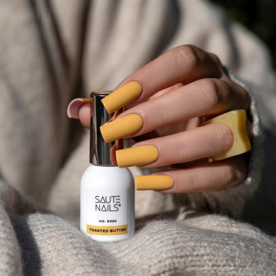 Unhas pintadas de amarelo seguram frasco de verniz branco SAUTE NAILS Toasted Butter.