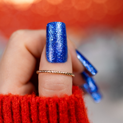 Unha longa com verniz azul glitter e anel dourado em fundo vermelho