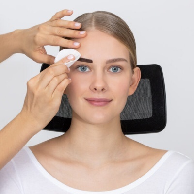 Mulher loira usa disco de algodão para limpeza da sobrancelha sentada numa cadeira preta