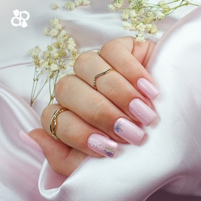 Unhas rosa claro com desenhos brancos e anéis dourados sobre tecido rosa e flores brancas