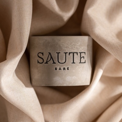 Frasco de cuidado pessoal cinza com rótulo SAUTE CARE sobre tecido bege