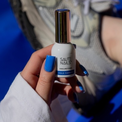Frasco de verniz para unhas branco com tampa metálica e texto, unhas azuis, ténis cinzentos ao fundo.