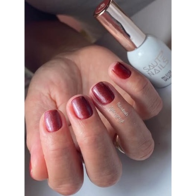 Mãos com unhas vermelhas metálicas e frasco de verniz branco com tampa dourada