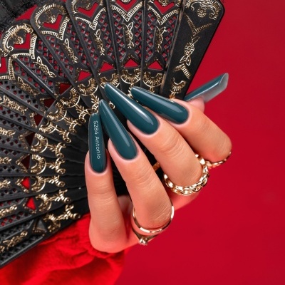 Mão com unhas verdes compridas segurando leque preto e dourado sobre fundo vermelho