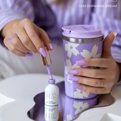 Mãos com unhas lilás a aplicar verniz SAUTE NAILS junto a copo térmico lilás com flores brancas