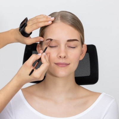 Mulher a ter sobrancelhas pintadas com pincel por mãos com pulseira preta, sentada em cadeira preta de rede, fundo branco