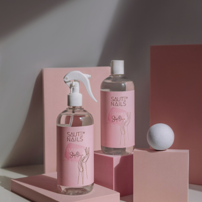 Dois frascos transparentes com rótulos rosa SAUTE NAILS sobre blocos rosa e uma esfera branca