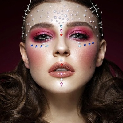 Mulher com maquilhagem rosa e adesivos de estrelas e pérolas no rosto, cabelo castanho ondulado