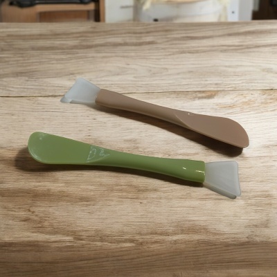 Dois utensílios de silicone verde e castanho claro sobre madeira clara