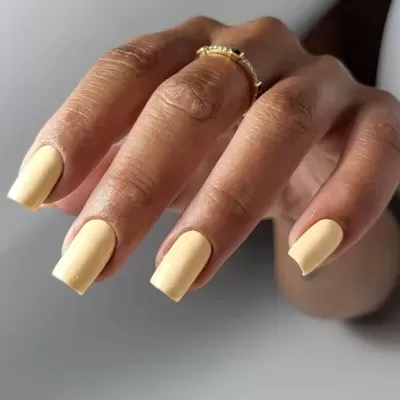Unhas pintadas de amarelo fosco com anel dourado em fundo cinza