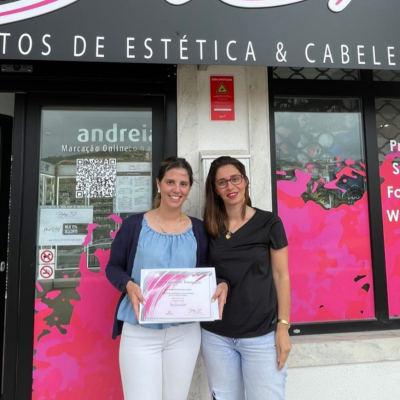 Duas mulheres em frente a uma loja de estética e cabeleireiro, uma segura um certificado