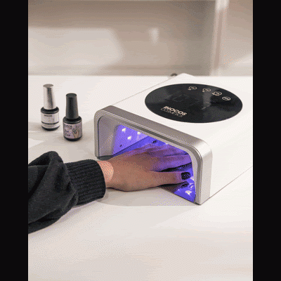 Secador de unhas LED branco a secar unhas femininas com dois frascos de verniz ao lado em mesa branca