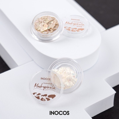 Duas embalagens pequenas de plástico com flocos de madrepérola, uma bege amarelada e outra branca, sobre fundo branco e preto, com etiquetas e marca INOCOS.