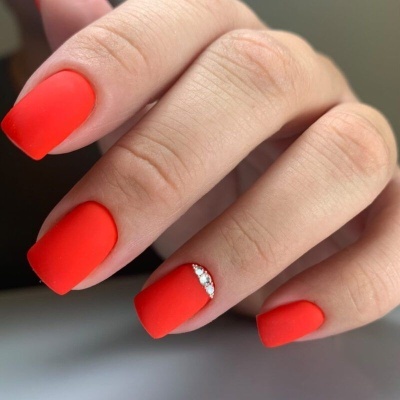 Unhas quadradas com verniz mate vermelho-laranja e detalhe de pedras brilhantes