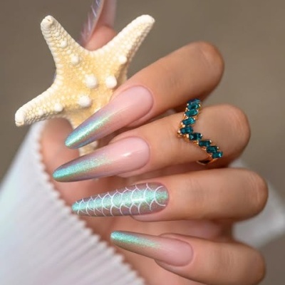 Mão com unhas stiletto holográficas rosa e verde, segurando estrela-do-mar branca, anel dourado com pedras azuis