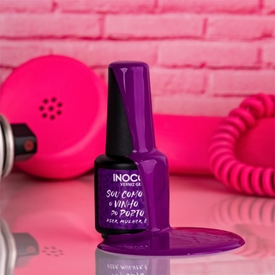 Frasco de verniz de unhas roxo com texto, verniz derramado sobre mesa espelhada, fundo rosa com parede de tijolos e telefone retro
