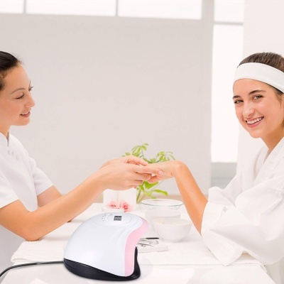 Dispositivo LED branco e rosa para unhas numa mesa branca com duas mulheres em ambiente luminoso