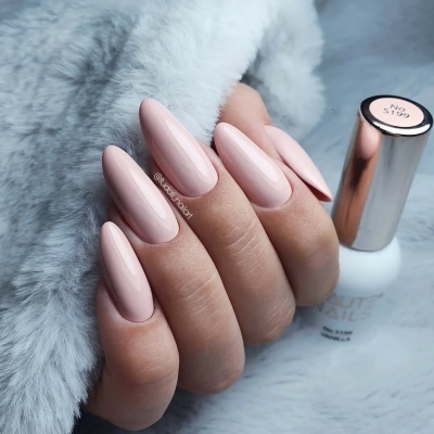 Unhas com verniz gel rosa claro em fundo de tecido cinzento felpudo com frasco de verniz branco e tampa dourada.