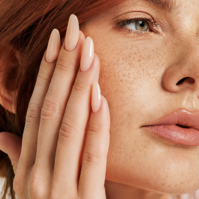 Close-up de mão feminina com unhas nude junto ao rosto com sardas