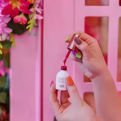 Mãos segurando frasco aberto de verniz SAUTE NAILS vermelho com fundo rosa e flores
