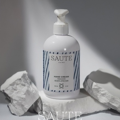 Frasco branco de creme para mãos SAUTE Care com doseador e etiqueta azul com riscas em fundo branco e pedras brancas.