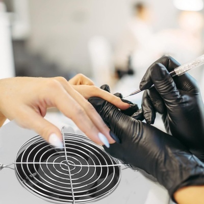 Manicure a aplicar produto nas unhas de uma pessoa com luvas pretas e bancada com grelha circular