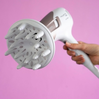 Secador de cabelo branco com difusor ligado segurado à mão sobre fundo rosa.