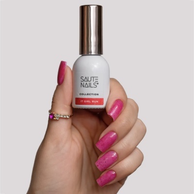 Frasco de verniz para unhas branco com tampa prateada e etiqueta vermelha, segurado por mão com unhas pintadas de rosa com pontos escuros