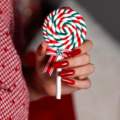 Mão com unhas vermelhas segurando pirulita colorida em espiral com laço vermelho e branco.