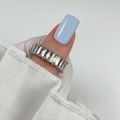 Anel prateado com pedras retangulares transparente no polegar com unha azul claro e tecido branco.