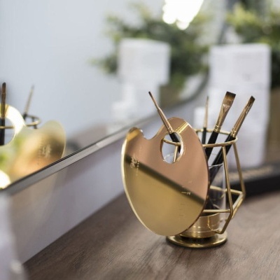 Paleta de pintura metálica dourada com suporte e pincéis em mesa de madeira junto a espelho