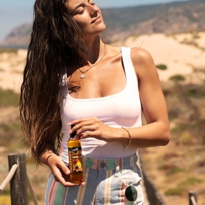 Mulher com top branco e calções às riscas a segurar garrafa com líquido âmbar na praia