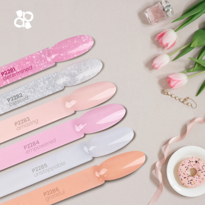 Amostras de verniz em tons pastel com etiquetas, flores, donut, perfume e fita rosa sobre fundo cinza.