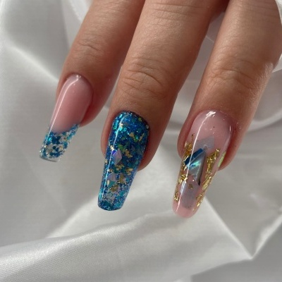 Unhas artificiais decoradas em azul, dourado e rosa claro com brilho e texturas sobre tecido branco