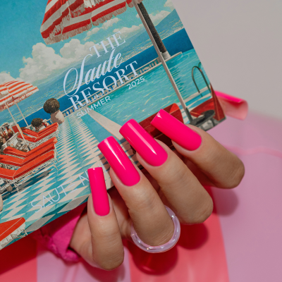 Mão com unhas rosa vibrantes segurando catálogo com imagem de resort e anel transparente.