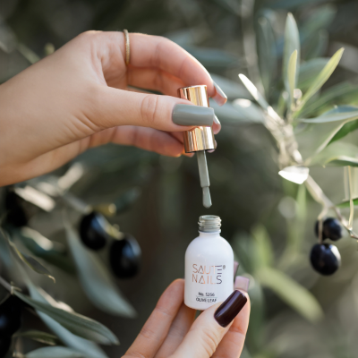 Verniz de unhas SAUTE NAILS branco com tampa dourada e mãos segurando-o junto a folhas de oliveira