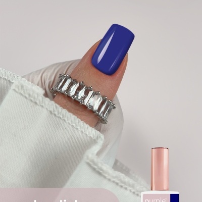 Unha com verniz gel azul escuro e anel prateado com pedras, tecido branco e embalagem de verniz gel