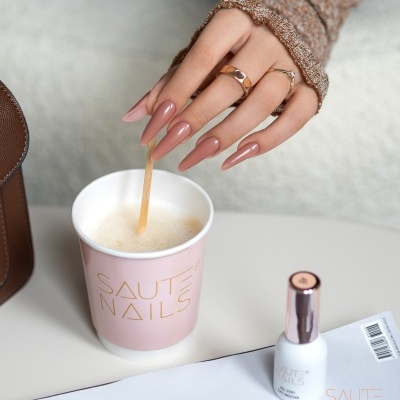Mão feminina com unhas longas nude rosadas segurando copo rosa SAUTE NAILS sobre mesa branca com esmalte e revista
