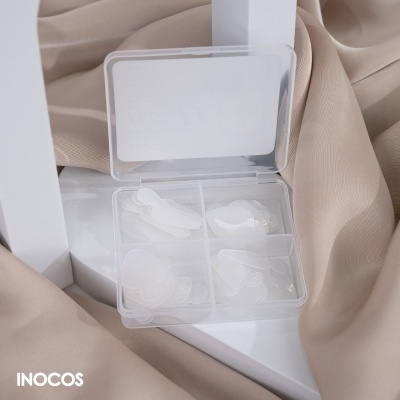 Estojo transparente com unhas postiças brancas em compartimentos, sobre tecido bege e superfície branca