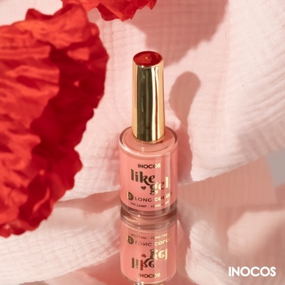 Frasco de verniz de unhas rosa claro com tampa dourada sobre superfície espelhada, com tecido branco e flor vermelha ao lado